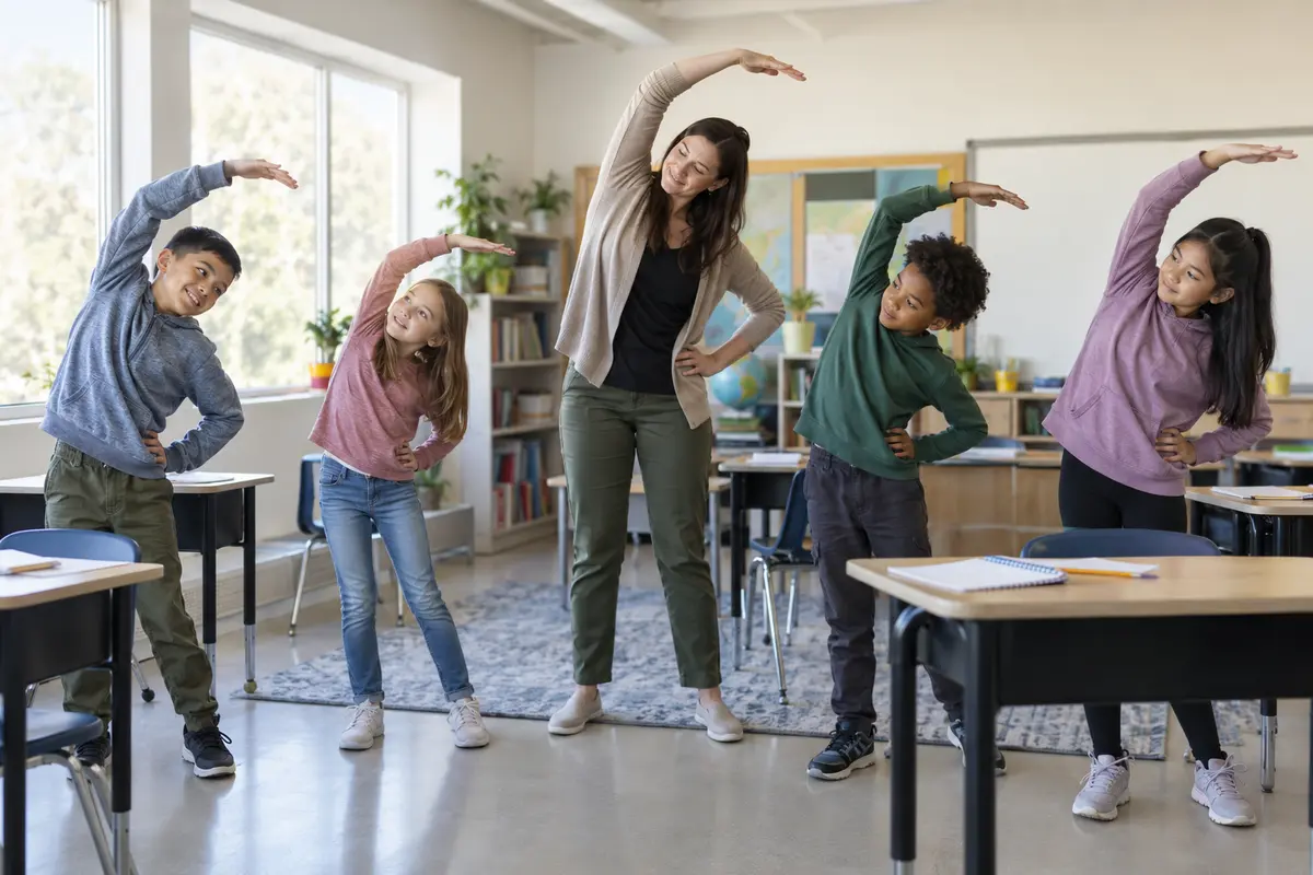 A teacher and four elementary students doing a gentle standing side stretch beside classroom desks.